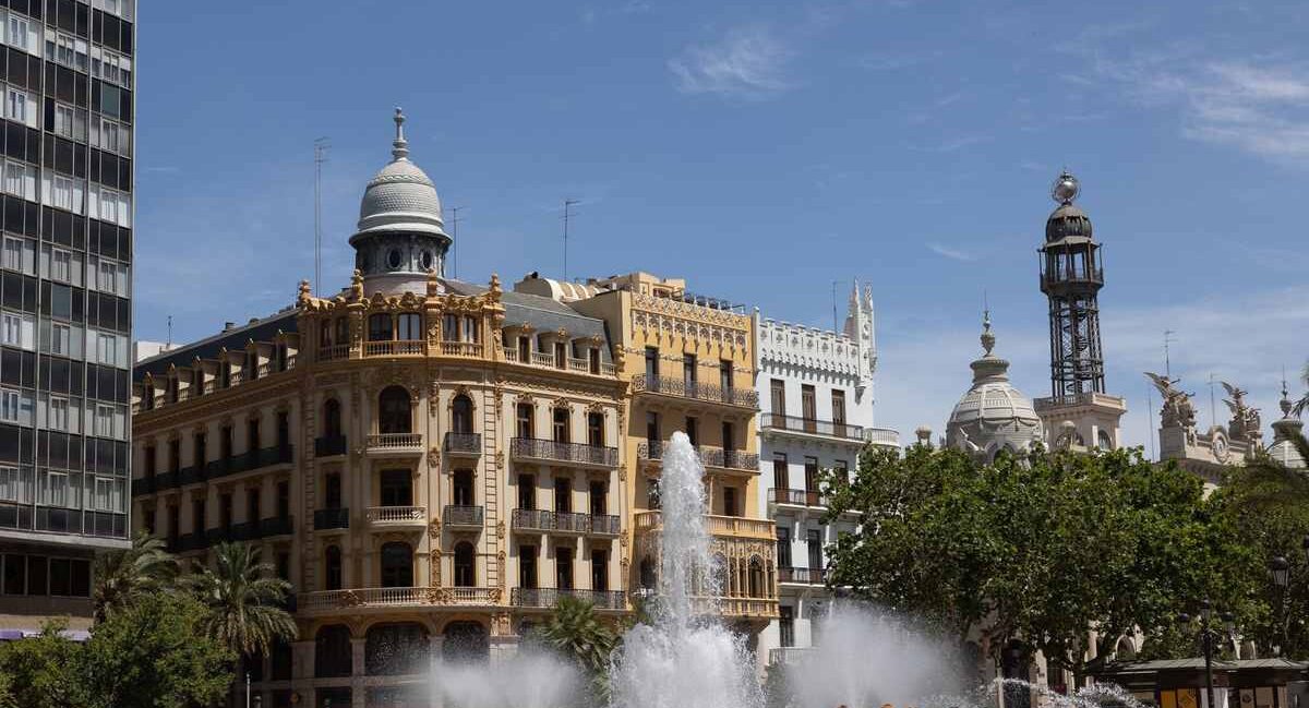 Un jet d'eau s'élève devant un bâtiment historique et une tour sous ciel clair