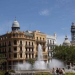Un jet d'eau s'élève devant un bâtiment historique et une tour sous ciel clair