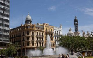Un jet d'eau s'élève devant un bâtiment historique et une tour sous ciel clair