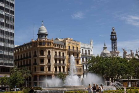 Un jet d'eau s'élève devant un bâtiment historique et une tour sous ciel clair