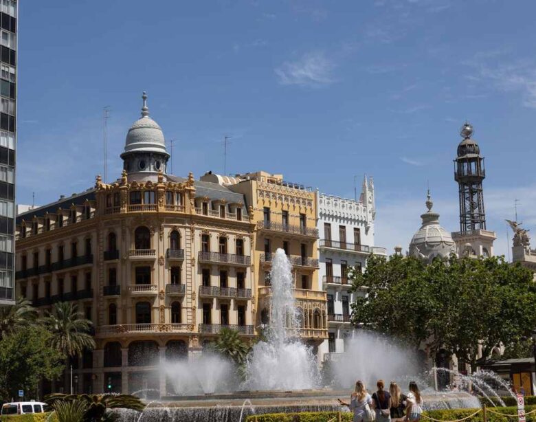 Un jet d'eau s'élève devant un bâtiment historique et une tour sous ciel clair
