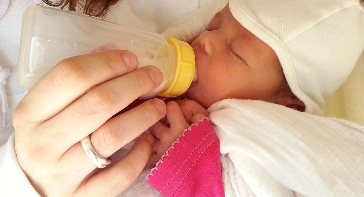 Une personne nourrit un nourrisson avec une biberette, chapeau blanc et manche rose